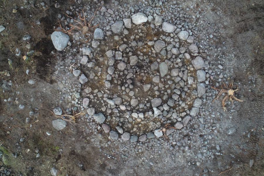 Stenformation arrangerad i cirkelmönster mot en jordig bakgrund. Stora och små stenar bildar flera sammanhängande cirklar, vilket antyder ett möjligt historiskt eller arkeologiskt fynd. Jorden runt stenarna är mörk och ojämn.