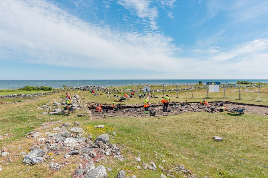 Arkeologer i orange och gula västar arbetar vid en utgrävning nära kusten. De undersöker en markyta omgiven av stenar och gräs. I bakgrunden syns havet och en klar himmel över Sandsby borg, Öland.