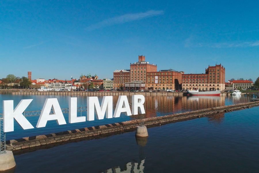 Stora vita bokstäver formar ordet "KALMAR" ovanför en pir vid vattnet. I bakgrunden syns gamla byggnader från Kalmar stad under en klarblå himmel, med speglingar i det lugna vattnet.