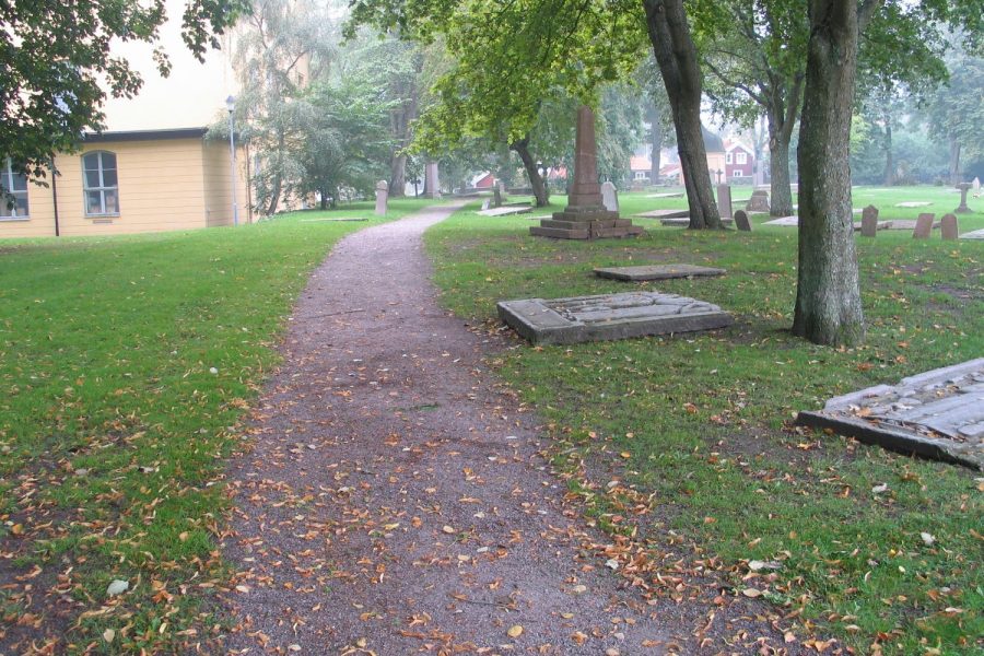 En grusgång slingrar genom Gamla Kyrkogården i Kalmar, omgiven av gamla gravstenar och höga träd. En gul byggnad i bakgrunden står mot en disig himmel. Löv täcker delvis marken.