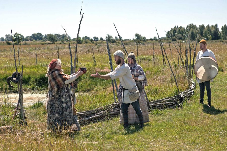 Fyra personer i historiska dräkter interagerar på en äng, med en man och en kvinna i förgrunden som delar en skål. Två andra personer står i bakgrunden, en med en stor trumma. Bakom dem syns enkla staket och ett landskap med träd.