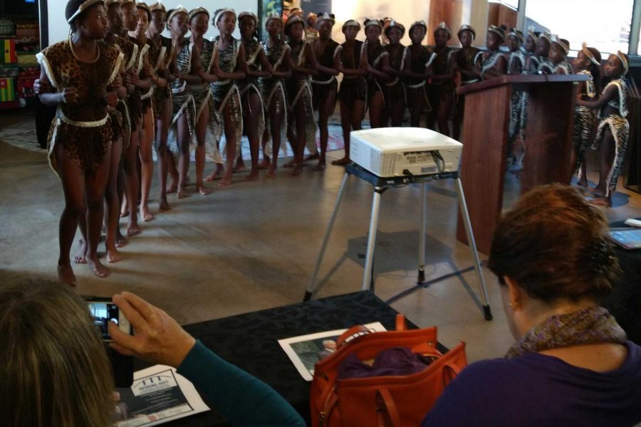 En grupp människor klädda i traditionella kläder dansar och sjunger framför ett podium under Bridging Ages-konferensen 2018 i Pretoria. Två sittande åskådare, en fotograferar och en annan tittar på. En projektor står nära podiet.