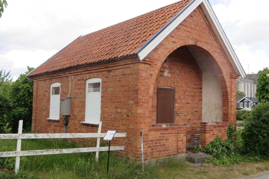 Rött tegelhus med spetsigt tak står vid en grusväg omgiven av grönska. Två små fönster syns på sidan, medan framsidan har en bågformad entré. I bakgrunden skymtas fler byggnader och blå himmel.