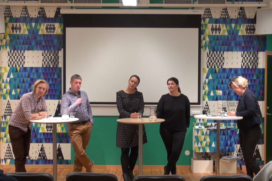 Fem personer deltar i en paneldiskussion i ett färgglatt rum, en man håller en mikrofon medan de andra lyssnar. Två personer antecknar. Bakgrunden pryds av ett geometriskt mönster i blått och grönt.