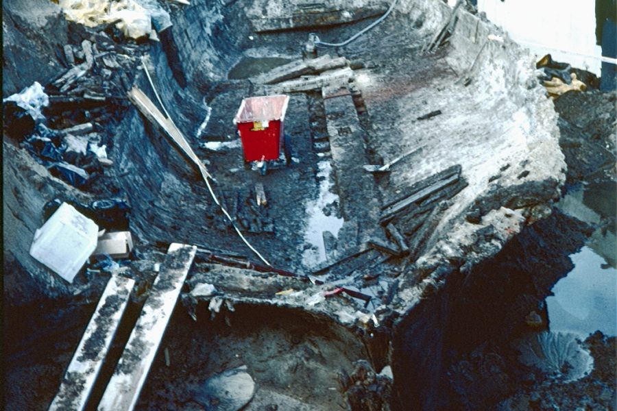 En djup utgrävning fylld med jord och vatten, med en röd tank i mitten. Omkring tanken syns betong, träbalkar och kablar. Området är uppenbart under byggnation eller renovering, med fokus på den centrala tanken.