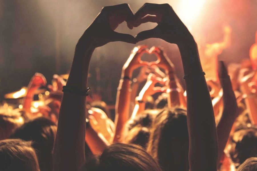 Person lyfter händer i en hjärtform mot en scen under en konsert. Flera händer syns i bakgrunden, alla bildar hjärtan, vilket skapar en känsla av gemenskap och glädje i den dunkla belysningen.