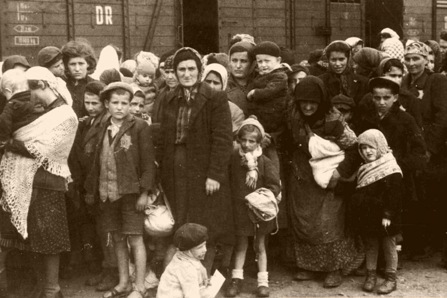 En grupp människor, inklusive barn och vuxna, står nära en godsvagn i en tågstation. De ser ut att ha få ägodelar och bär täta kläder. Situationen verkar spänd och omgiven av historisk kontext.