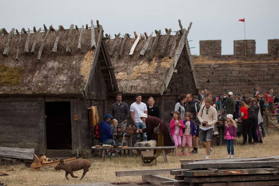 En gris springer framför en grupp människor nära gamla timmerhus med halmtak. Människorna, inklusive barn, samlas intresserat kring borden utanför byggnaderna. I bakgrunden syns en stenmur och en röd flagga.