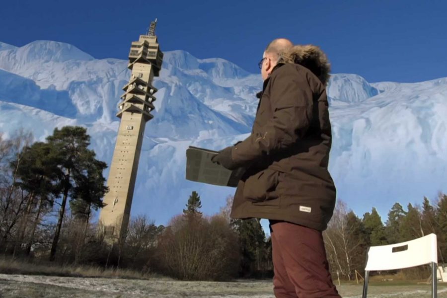En person i vinterjacka står utomhus med en bärbar dator i händerna, framför ett högt telekommunikationstorn med snötäckta berg i bakgrunden. Soligt väder belyser naturen och den vita stolen bredvid.