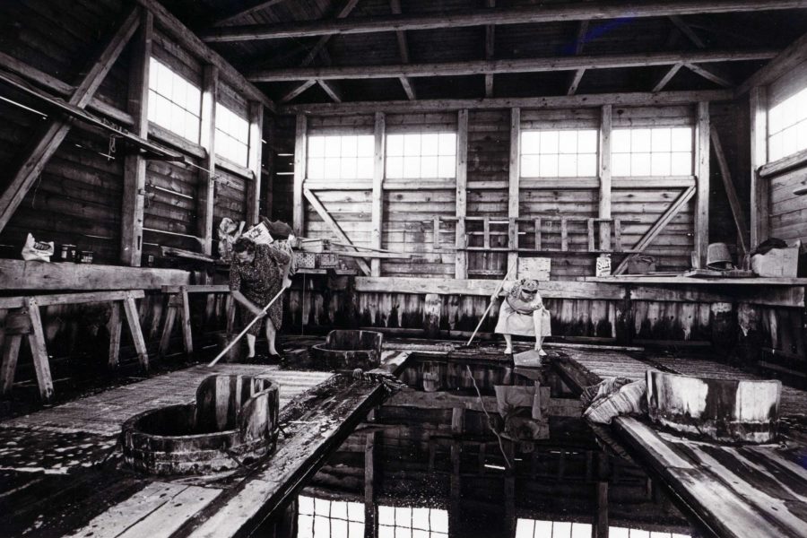 Två personer arbetar i ett gammalt trähus med högt i tak, belyst av fönster. De håller långa träspadar och rör om i stora träkar fyllda med vatten. Väggarna har hyllor och verktyg, vilket skapar en rustik atmosfär.