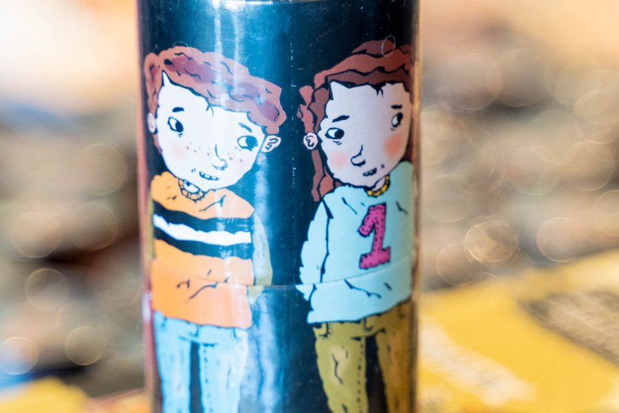 En burk med illustration av två tecknade barn. Barnen står bredvid varandra på en svart bakgrund. Burken står på en bok med en tecknad stadsmiljö. Färgerna är livfulla och kontrasterande.