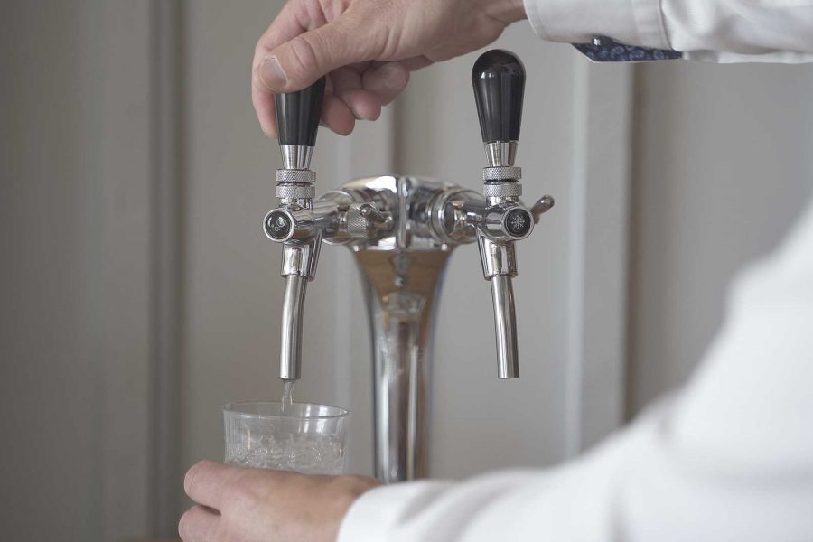 En person håller ett glas under en tvågrepps vattenpump och fyller det med vatten. Pumpen har två svarta handtag med symboler som indikerar olika vatteninställningar, monterad på en metallisk bakgrund.