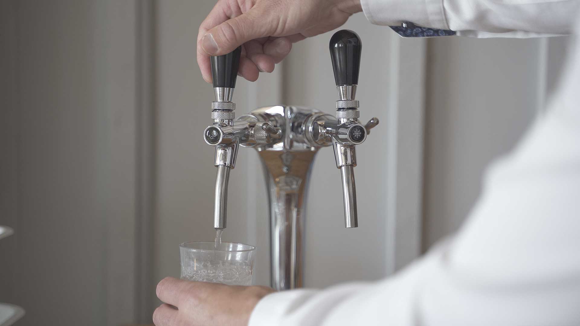 En person håller ett glas under en tvågrepps vattenpump och fyller det med vatten. Pumpen har två svarta handtag med symboler som indikerar olika vatteninställningar, monterad på en metallisk bakgrund.