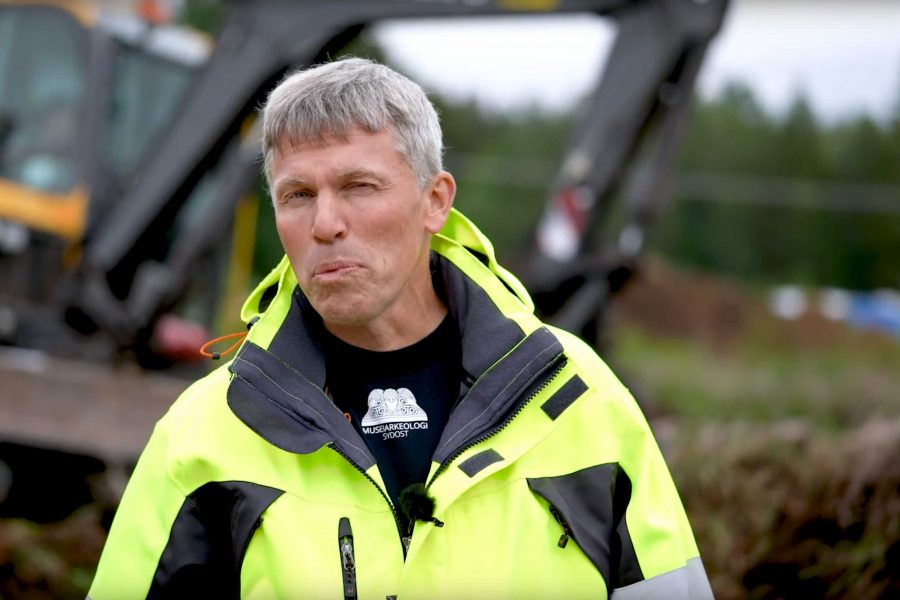 En man i gul jacka står framför en suddig bakgrund med en grävmaskin. Han bär en svart tröja med texten "MUSARKOLOGI SYDOST". Han har kort, grått hår och ser ut att vara utomhus vid en arbetsplats.