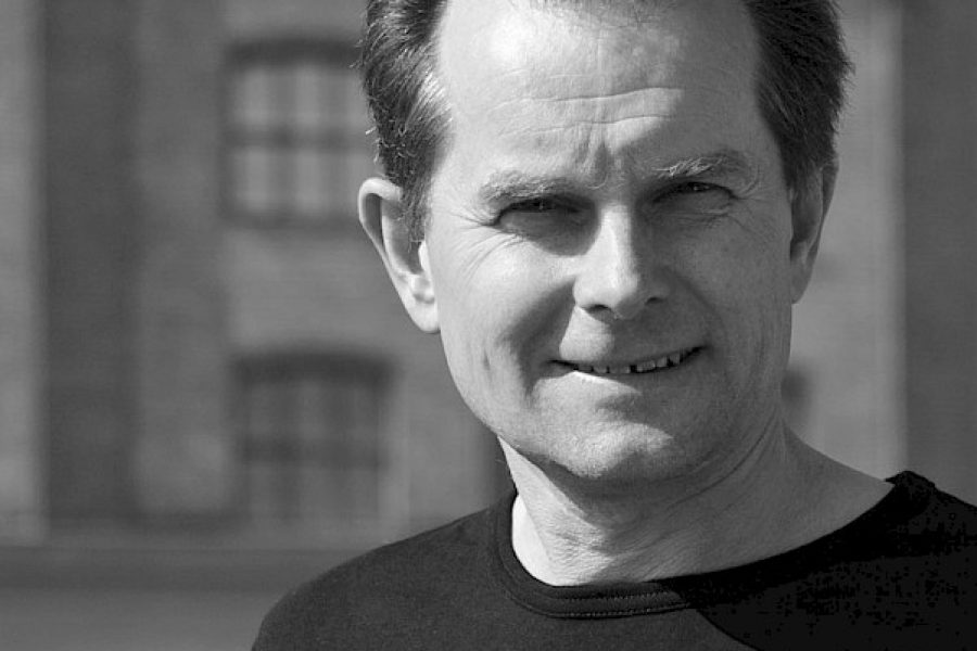 Man i medelåldern med kort hår bär en mörk tröja och ler mot kameran. Han står framför en suddig byggnad vilket tyder på en utomhusmiljö. Bilden är i svartvitt och han ser avslappnad ut.