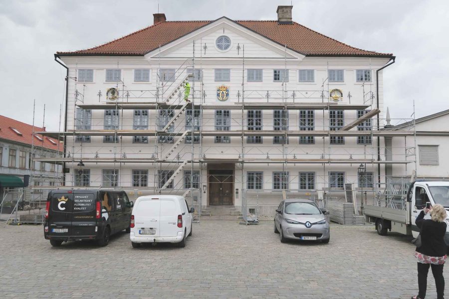 Rådhuset i Kalmar står bakom byggnadsställningar för renovering. Tre bilar är parkerade framför, och en person fotograferar. Byggnaden har ett rött tegeltak och ett vapensköld på fasaden.