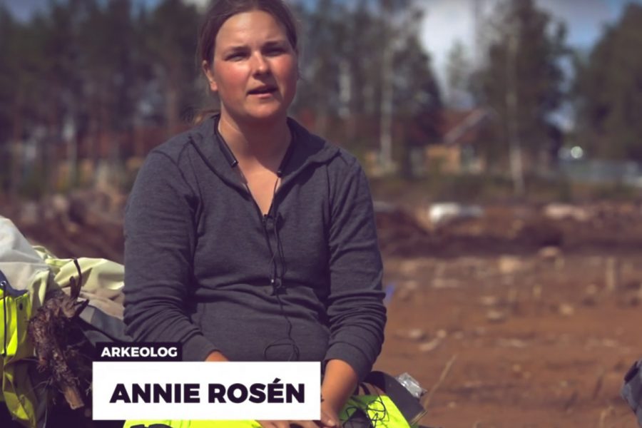 Arkeolog Annie Rosén sitter utomhus på en arkeologisk utgrävning, omgiven av jord och träd i bakgrunden. Hon bär en grå tröja och gula byxor. Text på bilden: "Arkeologi Annie Rosén". Text ovan: "Arkeologidagen 2019".
