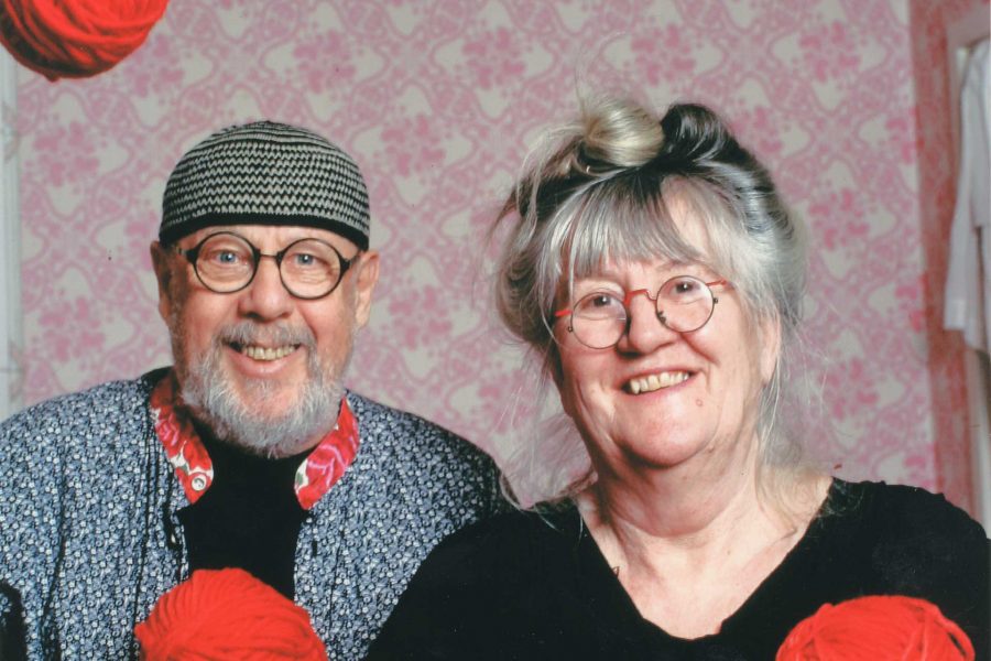 Ett äldre par ler mot kameran i ett rum med rosa tapet. Mannen håller ett rött garnnystan och bär en mössa och glasögon, medan kvinnan också håller i garn och har håret uppsatt.