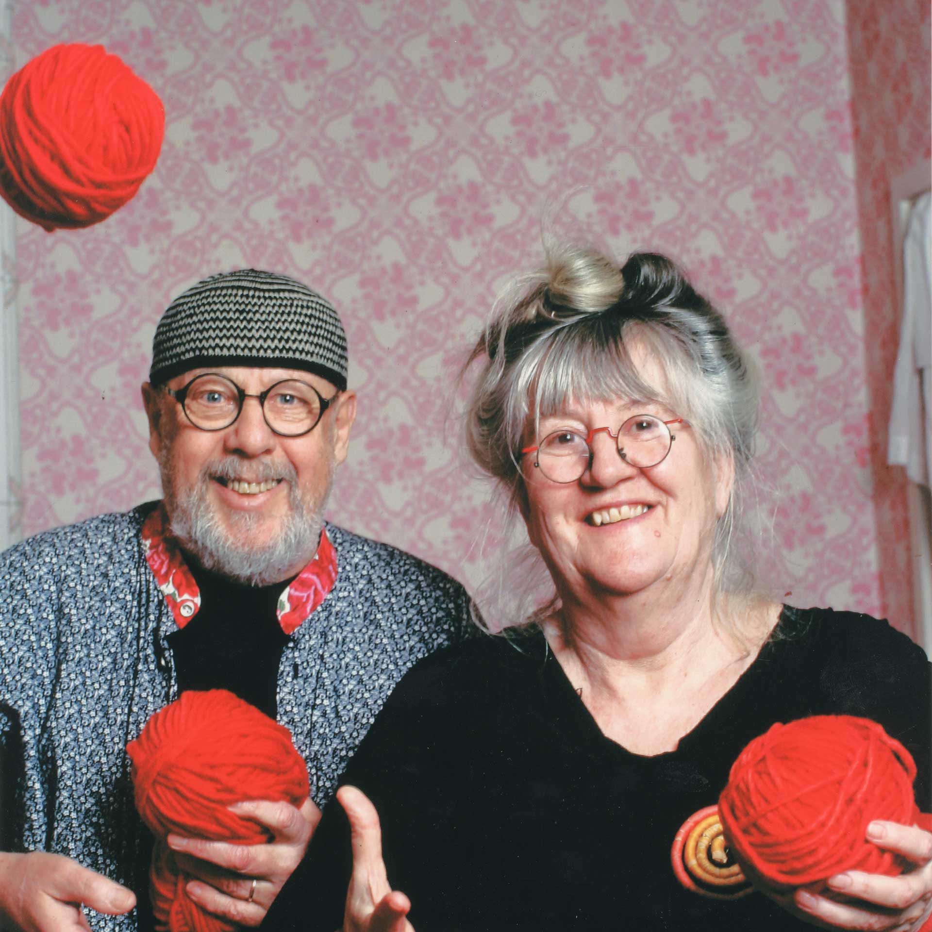 Ett äldre par ler mot kameran i ett rum med rosa tapet. Mannen håller ett rött garnnystan och bär en mössa och glasögon, medan kvinnan också håller i garn och har håret uppsatt.