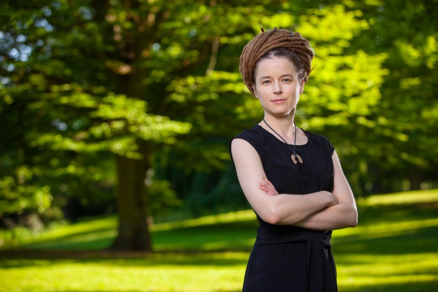 En person med hår uppsatt i långa dreadlocks står med armarna i kors i en grön park. Hon bär en svart ärmlös klänning och ett halsband med trädetaljer. Detaljerade träd och gräsmatta syns i bakgrunden.