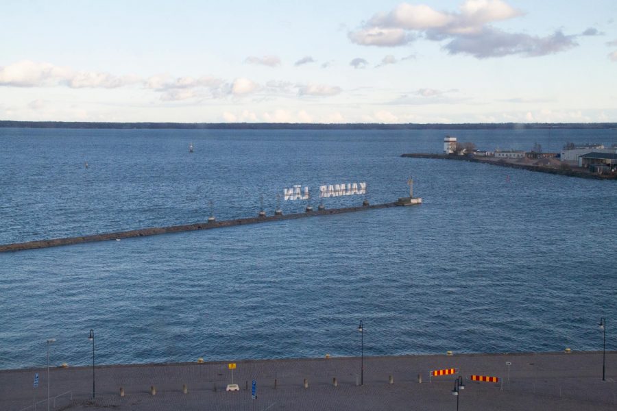 En vy över en hamn med en lång pir som sträcker sig ut i havet. På piren finns en stor text med bokstäverna "KALMAR LÄN". I förgrunden syns en stenlagd yta och parkeringsskyltar.