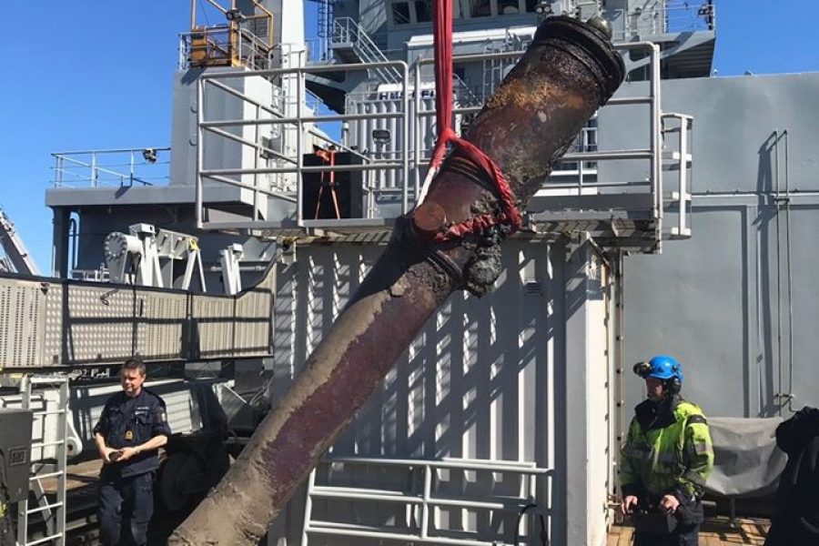 En stor kanon lyfts ned på ett fartygsdäck med hjälp av två röda remmar. Två personer, en i uniform och en i säkerhetsutrustning, övervakar processen. Bakgrunden visar fartygsstrukturer under en klarblå himmel.