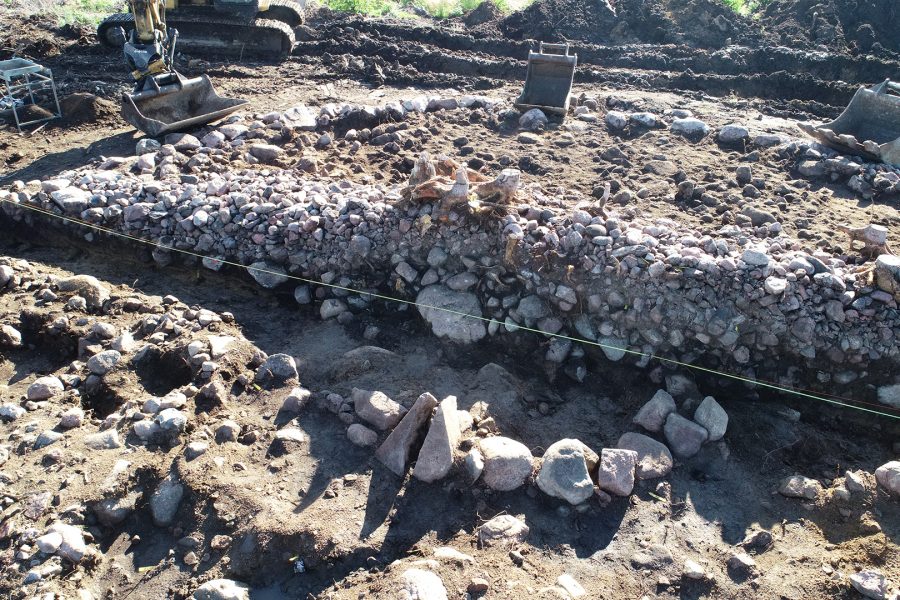 En grävmaskin arbetar på en arkeologisk utgrävningsplats, där en mur av stenar och jord har blottlagts. Solen skiner på området, och en linje av rep markerar utgrävningens gränser.