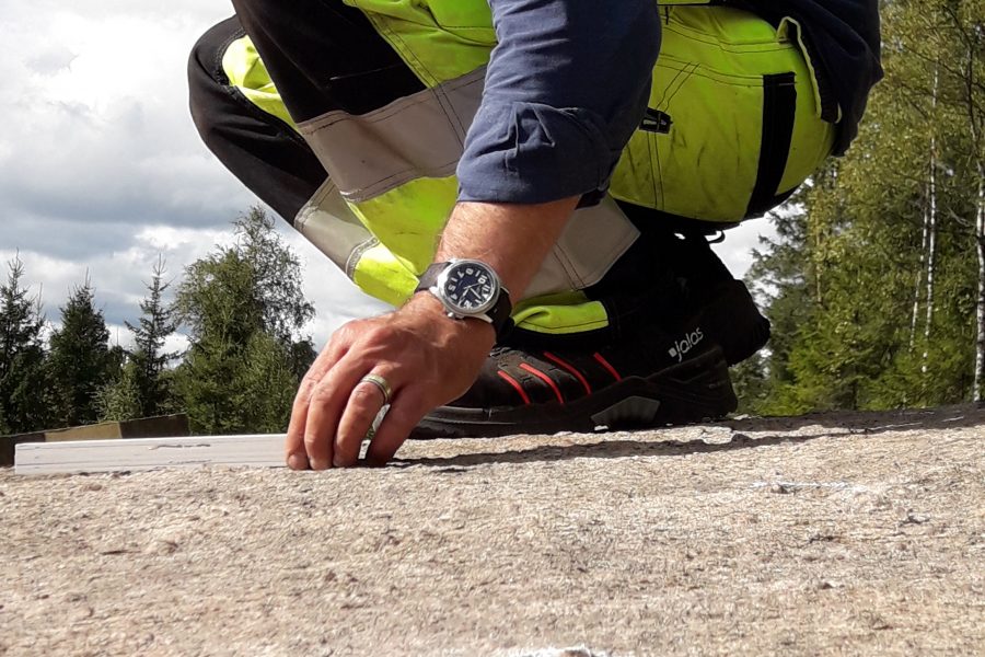 En person iförd gul skyddsbyxa och blå skjorta sitter på huk på en stenig yta, håller en kamera. Handen vilar på marken nära en vit mätsticka. Skog och molnig himmel syns i bakgrunden.