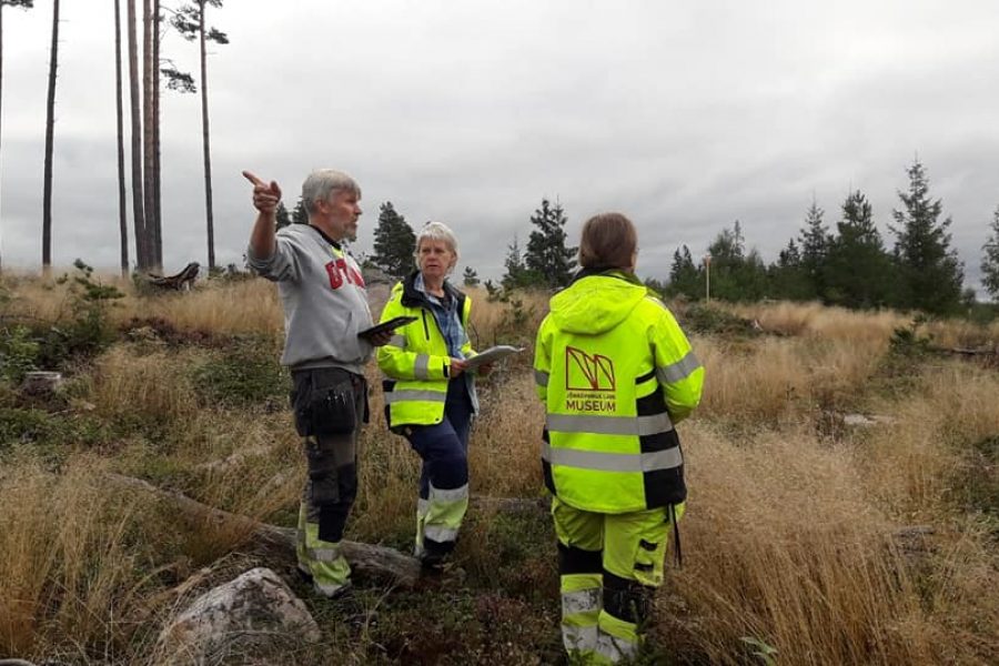 Tre personer diskuterar i ett öppet landskap med högt gräs och skog i bakgrunden. Två bär varselkläder med texten "Museum". En man pekar mot horisonten medan en kvinna håller i ett block.