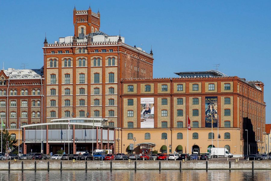 En tegelbyggnad med flera våningar, Kalmar läns museum, står vid vattnet. Två stora affischer pryder fasaden, en med texten "Kalmar läns museum". Bilar parkeras framför byggnaden under en klarblå himmel.