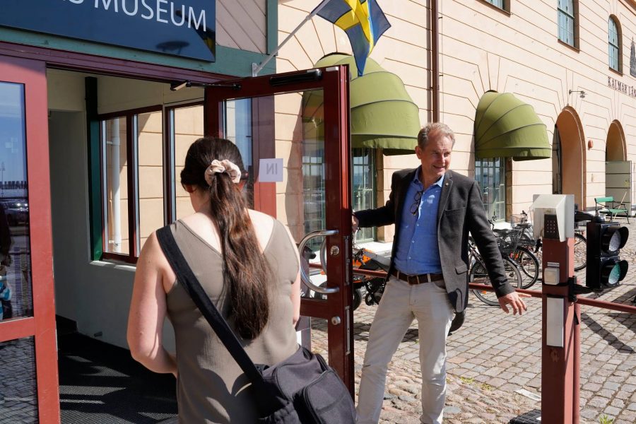 En man öppnar dörren till Läns Museum för en kvinna som är på väg in i byggnaden. En svensk flagga vajar ovanför ingången, och flera cyklar är parkerade utanför. Solsken belyser scenen.