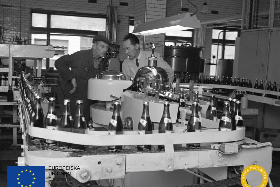 Två män samtalar vid en flaskproduktion i en fabrik. Flaskorna transporteras på ett löpande band. I bilden syns logotyper för Europeiska unionen och länsstyrelsen i Kalmar län. Miljön är industriell.