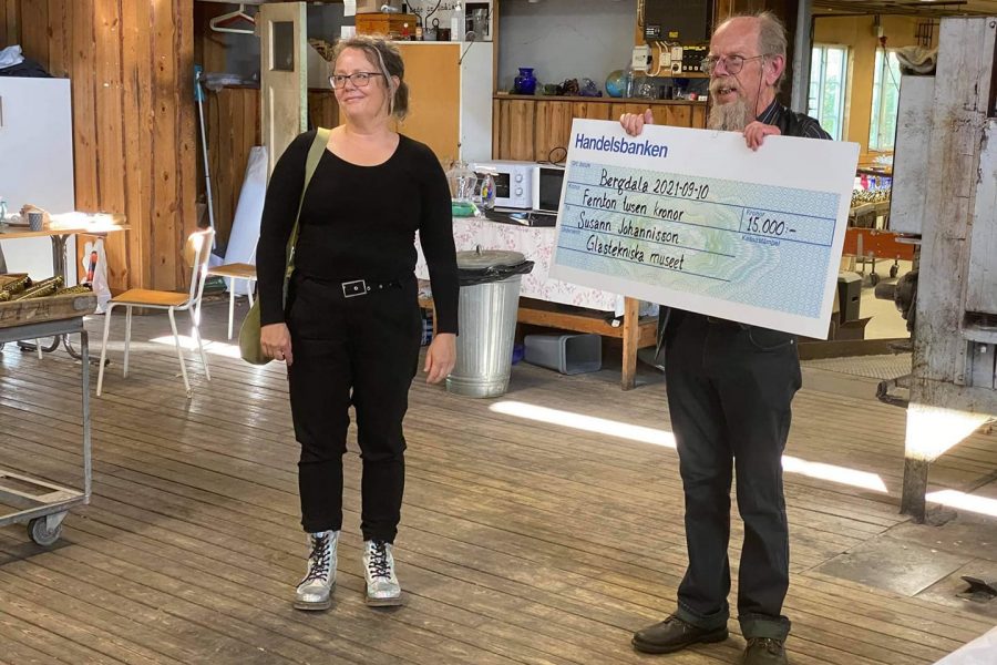 Susann Johannisson står bredvid en man som håller en stor check. Checken är från Handelsbanken och daterad Bergdala 2021-09-10 för femton tusen kronor till Susann Johannisson, Glastekniska museet. Rummet har träväggar och trägolv.