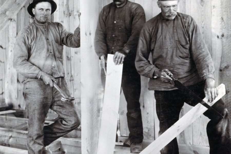 Tre män arbetar på ett husbygge i Kråksmåla, klädda i tidstypiska kläder. En man håller en hammare, medan de andra två sågar en träplanka. Bakgrunden visar en vägg av träpaneler, vilket ger en historisk känsla.