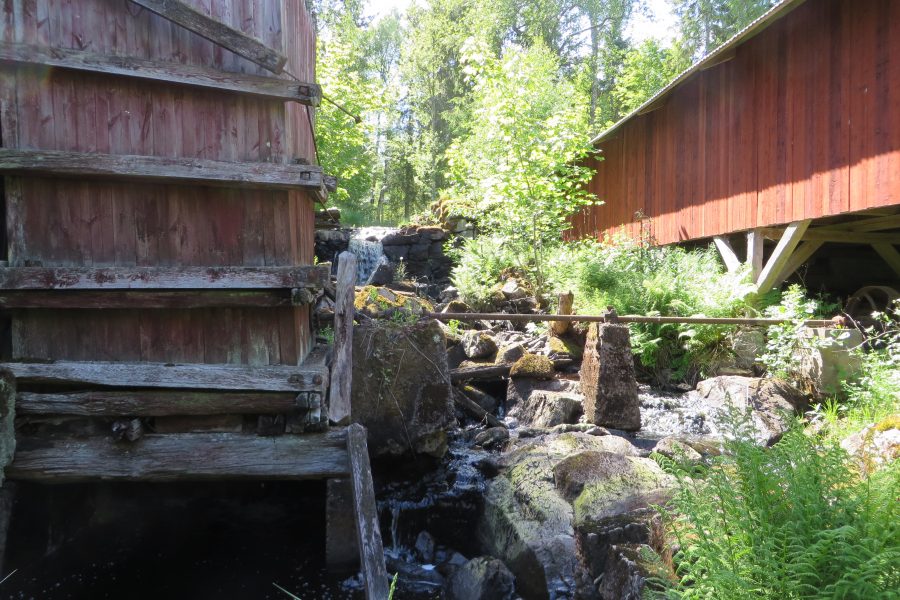 Gammalt trähus intill en liten fors, omgiven av grönska och solbelysta träd. Vatten rinner över stenar nedanför bron. Övergivet hjul och mossa täcker platsen, vilket skapar en känsla av lugn i skogen.