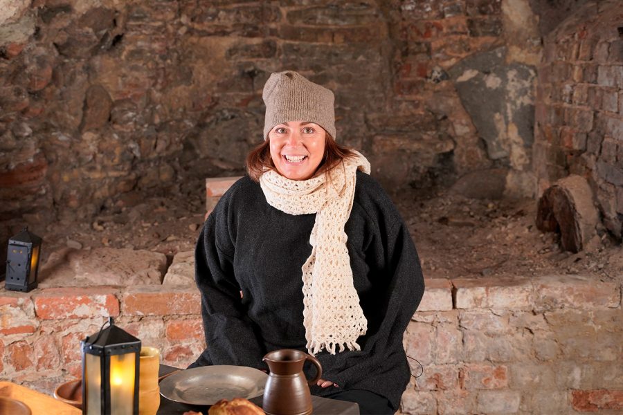 Kvinna klädd i mörk tröja, beige mössa och vit halsduk, ler framför en gammal tegelvägg. Framför henne finns ett bord med en metallmugg, tallrik och en lykta som ger ett varmt ljus.