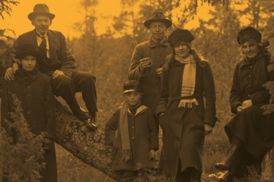 En grupp på sex personer, klädda i varma kläder och hattar, poserar i skogsmiljö. Två vuxna sitter på ett träd medan övriga står bredvid. En person håller i en kopp, medan andra bär halsdukar.