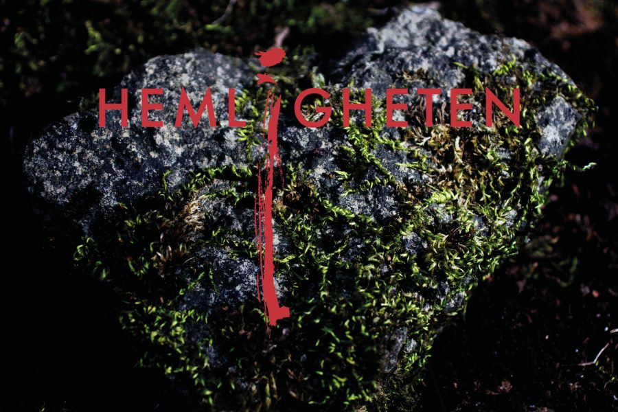 Mossbelagd hjärtformad sten med texten "HEMLIGHETEN" i rött ovanpå. Ett vertikalt rött streck betonar bokstaven "I". Stenen ligger på mörk, fuktig mark, vilket ger en mystisk känsla.