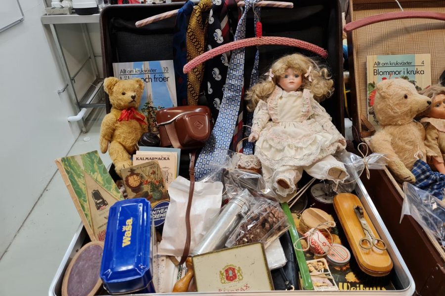 Ett öppet resväska fyllt med olika föremål: en docka i spetsklänning, flera nallebjörnar, böcker om krukväxter, slipsar, en blå Wasa-burk, skönhetsprodukter och souvenirer. Resväskan står i en rum med andra väskor och vintageobjekt.