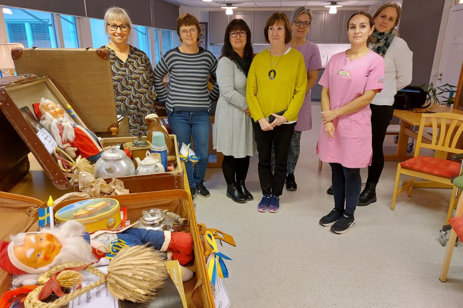 Sju personer stående i ett rum, varav tre i färgglada kläder. Framför dem finns en öppen koffert fylld med traditionella svenskrelaterade föremål, inklusive en jultomtefigur, flaggor och gamla leksaker, vilket skapar en nostalgisk stämning.