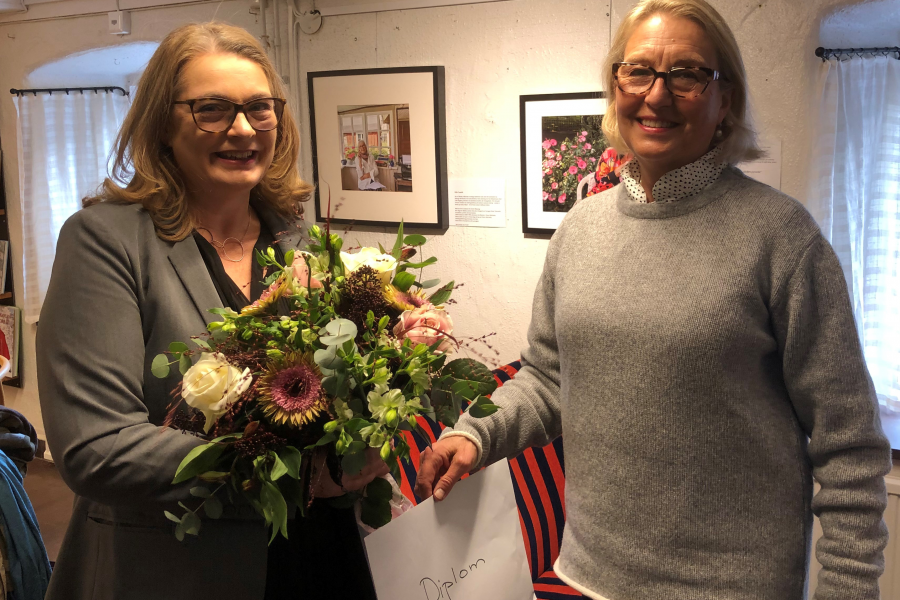 Två kvinnor ler i ett rum med tavlor på väggarna. Den ena håller en stor blombukett och den andra ett kuvert märkt "Diplom". Miljön ser ut som ett galleri med ljusa väggar.