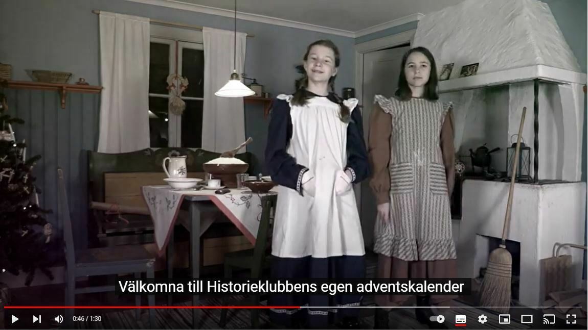 Två flickor i förkläden står framför en spis i ett gammaldags inrett kök. En julgran syns till vänster och ett dukat bord med kanna och fat. Text: "Välkomna till Historieklubbens egen adventskalender".