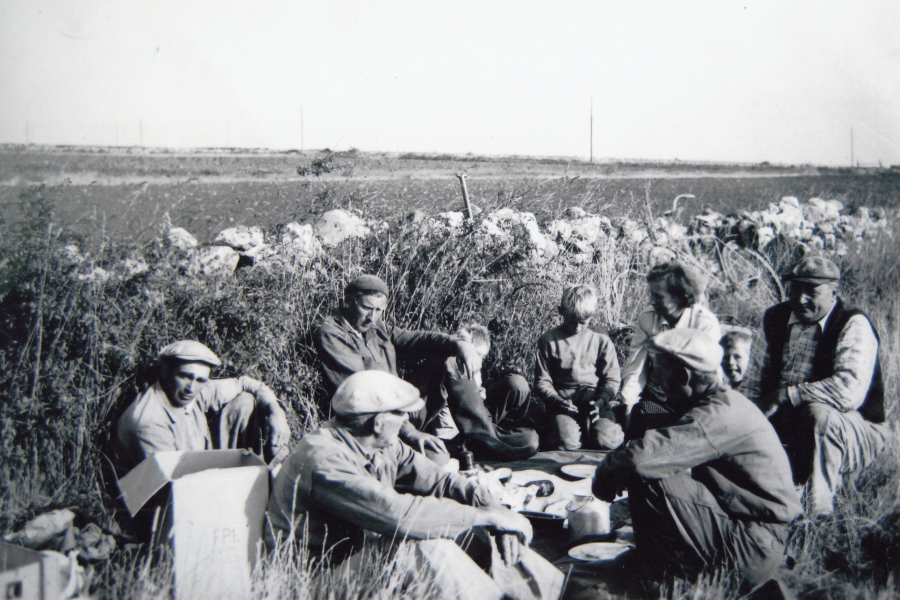 En grupp människor sitter tillsammans och har fika i en lantlig miljö, omgivna av en stenmur och fält. Flera vuxna och barn deltar i samlingen. Det verkar vara en paus från jordbruksarbete eller liknande daglig aktivitet.