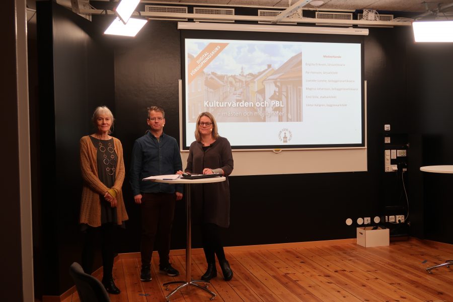 Tre personer står framför en presentationsskärm i ett mötesrum. Skärmen visar texten "Kulturvärden och PBL – måsten och möjligheter" samt namn på medverkande, inklusive Birgitta Eriksson, Per Hansson och Liselotte Jumme. Rummet är modernt inrett.