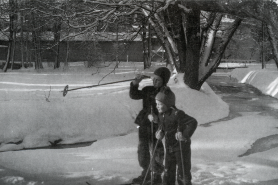 Två barn åker skidor på snö vid en frusen bäck, omgivna av snötäckta träd och byggnader i bakgrunden. Det främre barnet pekar framåt med en stav medan det andra följer efter.