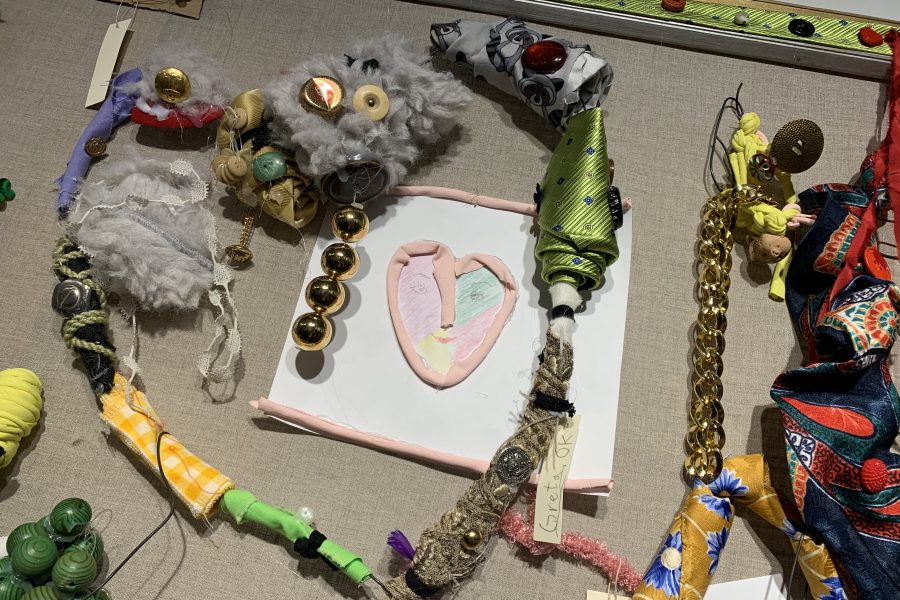 Färgglada textilobjekt med knappar, tyg och kedjor är kreativt arrangerade på en tavla. I mitten finns ett hjärta i lera med målad textur. Märkning "Greta" syns på en av objekten.