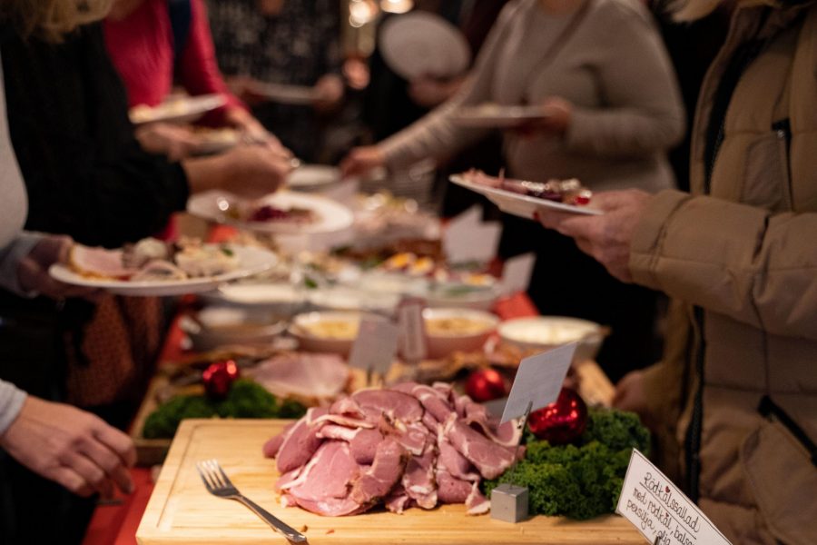 Gäster serverar sig själva vid en buffé med julmat. Ett bord är täckt med olika rätter som skinka och rödkålssallad, dekorerat med grönsaker och röda julprydnader. Flera personer står i kö med tallrikar.
