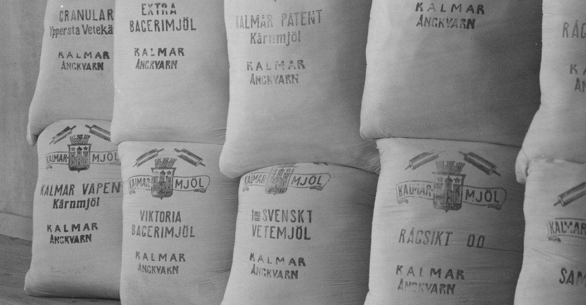 Säckar av mjöl står staplade i tre rader mot en ljus vägg. Varumärken och text på säckarna inkluderar "Kalmar Ångkvarn" och olika mjölsorter såsom vetemjöl, kärnmjöl och rågsikt. Golvet är av trä.