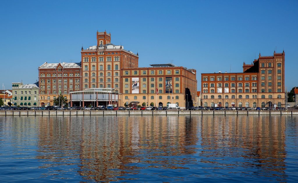 Stort tegelbrukskomplex speglas i vattnet under en klarblå himmel. De olika byggnaderna har detaljerad arkitektur med valv och torn. Flera reklamaffischer hänger på fasaderna, och bilar parkerade längs kajen kompletterar vyn.