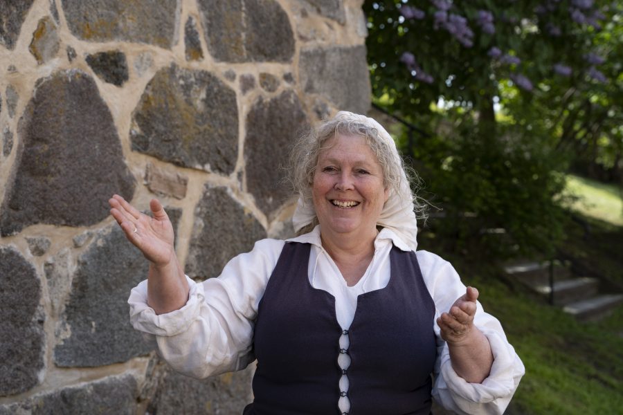 Kvinna klädd i traditionella kläder gestikulerar glädjefyllt framför en stenmur. Bakgrunden består av gröna buskar och syrenbuskar med blommor. Solsken belyser scenen, vilket skapar en varm och vänlig atmosfär.